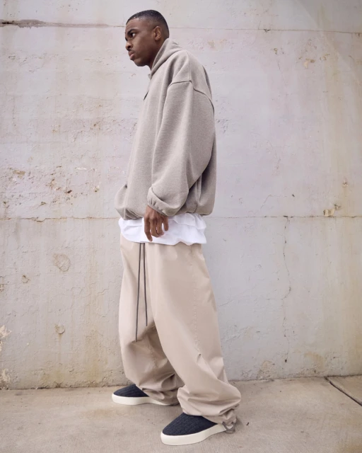 Man in beige oversized sweater and pants stands against a concrete wall, showcasing a casual yet stylish outfit and urban aesthetic.