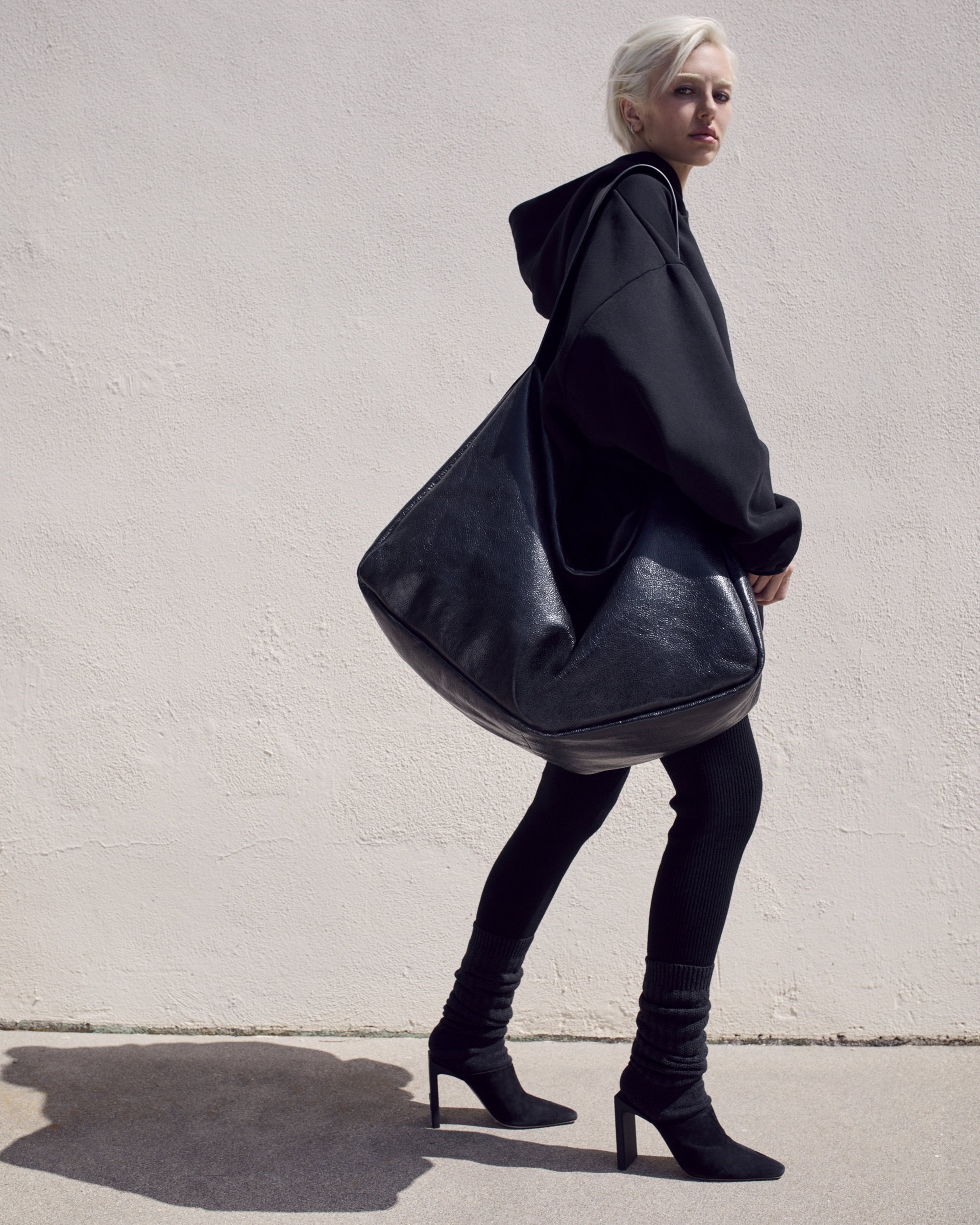 A woman in a black hoodie and leggings carries a large black leather bag, walking against a neutral wall in a fashion shot.