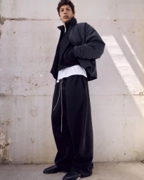 Model standing against a concrete wall wearing a dark puffer jacket, layered white t-shirt, and relaxed black trousers.