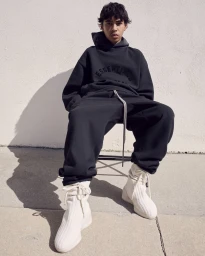 A young male model seated on a chair wearing a dark hoodie, matching sweatpants, and cream-colored high-top sneakers.