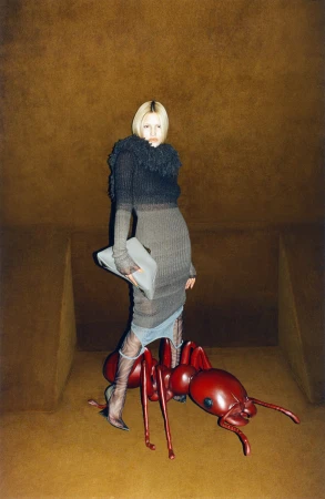 A fashionable woman in a grey dress and black top poses next to a large red ant in an oddly lit, neutral-toned studio-like setting.