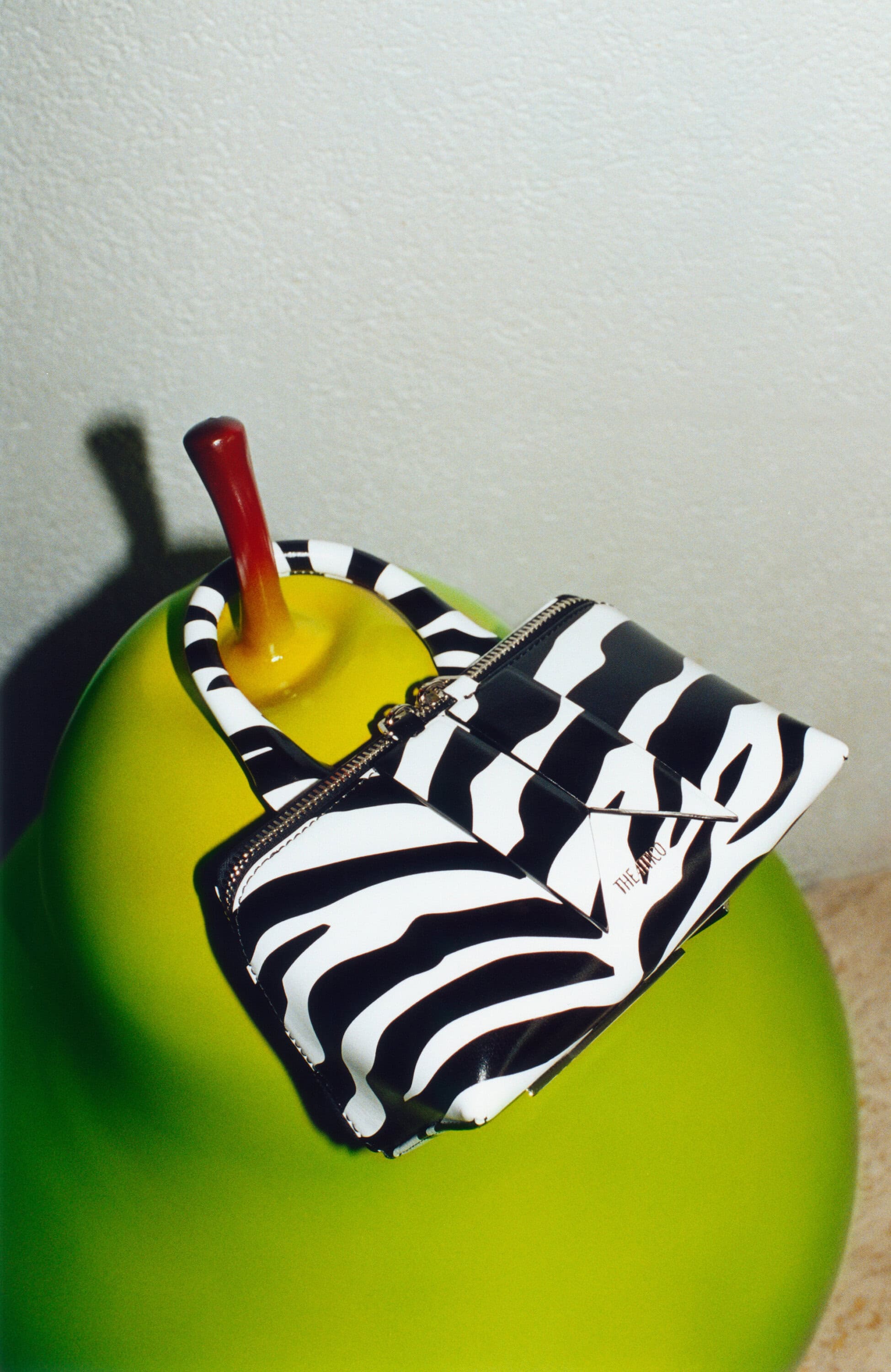 Zebra print purse on a glossy green pear-shaped object against a light grey wall and surface, showcasing fashion accessories.