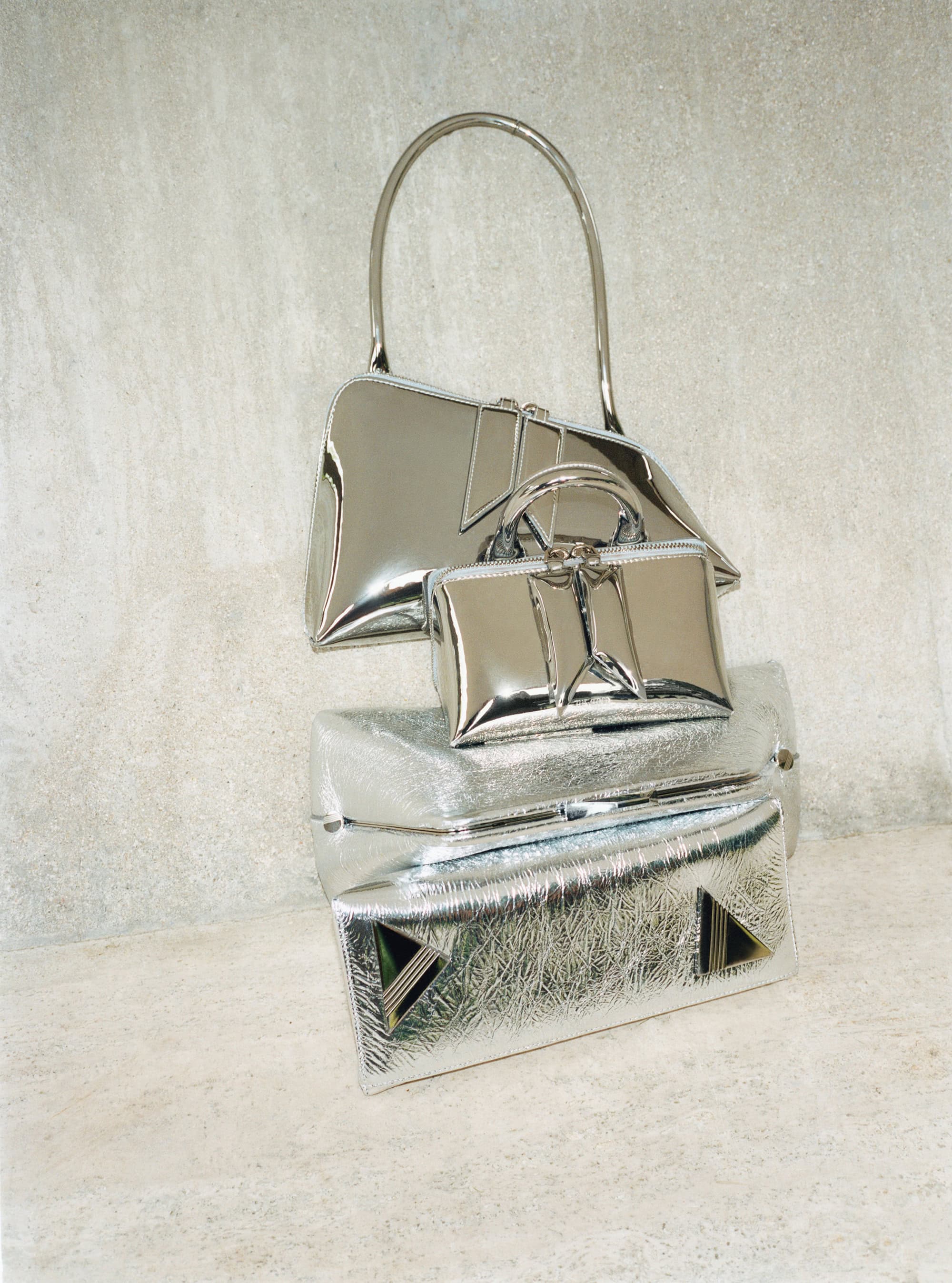 A stack of three shiny silver purses of varying sizes, including a handbag and two clutch bags, are displayed indoors.