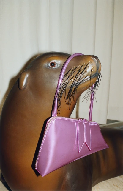 Lilac satin purse with a bow detail is displayed balanced on the snout of a brown decorative seal in a brightly lit indoor scene.