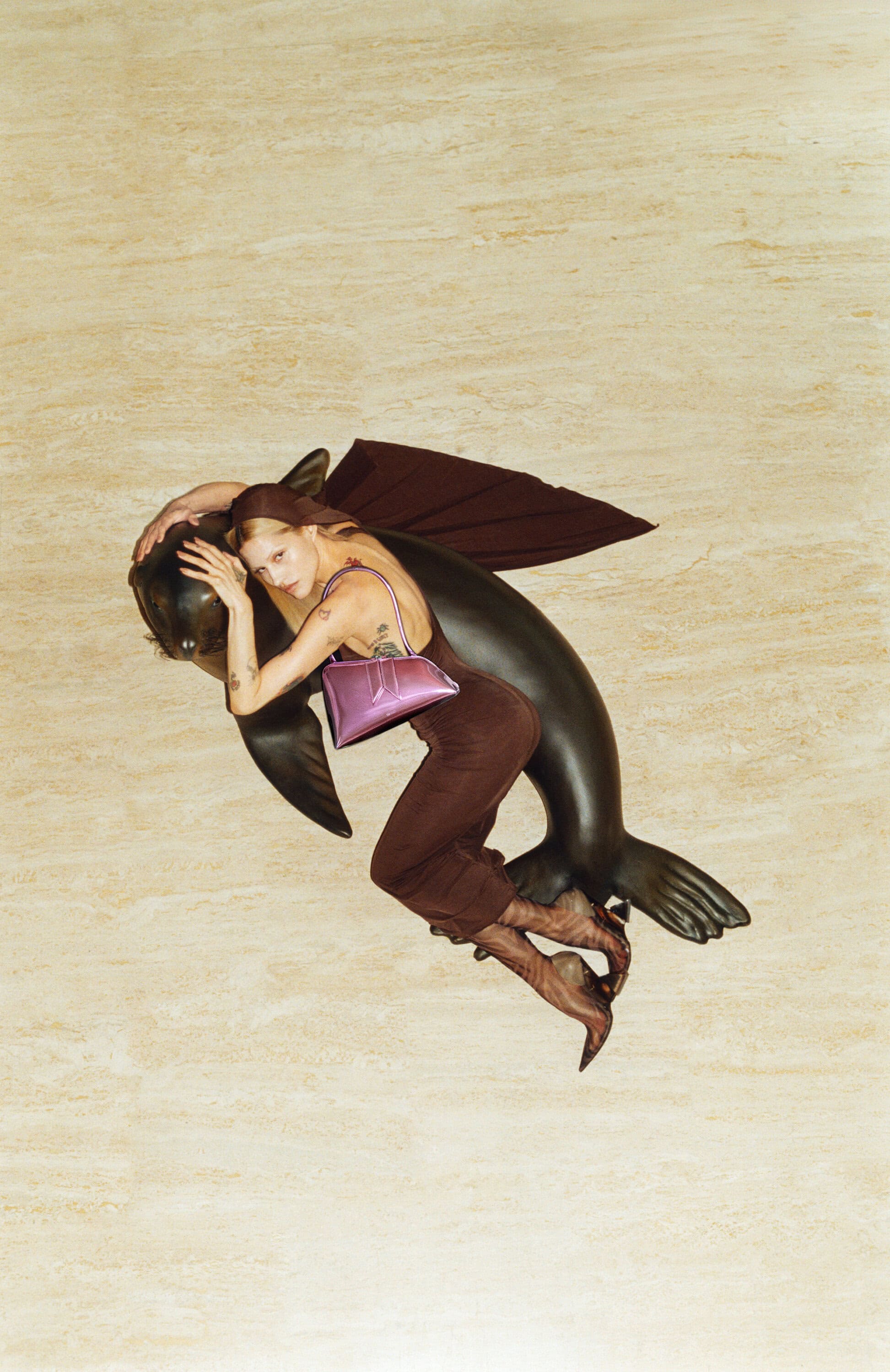 A fashion photo features an adult female model in brown, posing with a black fish statue against a tan wall, wearing a pink purse.