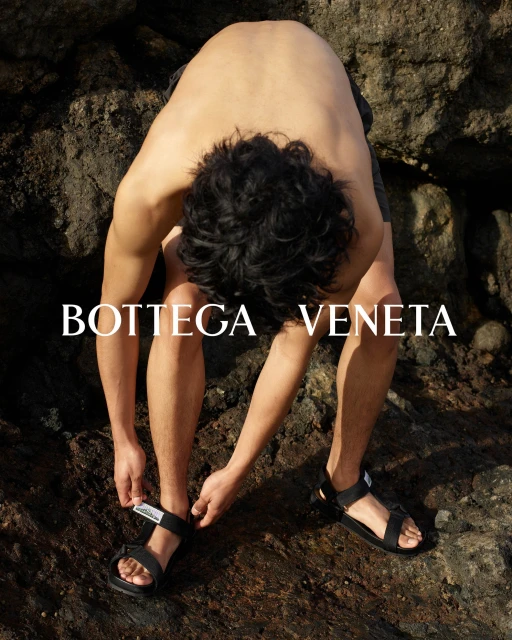 A tan adult male adjusts the strap of his black sandal while sitting on a rock; outdoor fashion advertisement for Bottega Veneta.