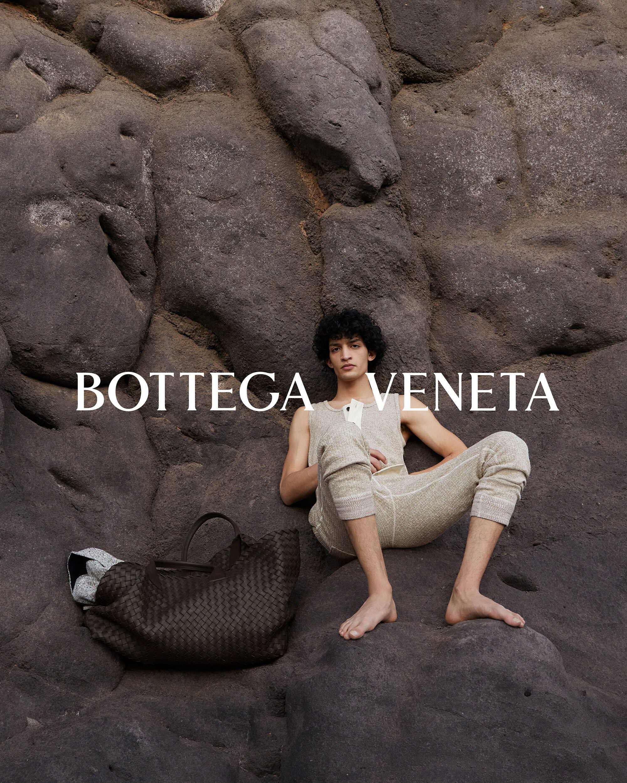 A man reclining barefoot against a rocky stone surface next to a dark woven leather tote bag for Bottega Veneta.