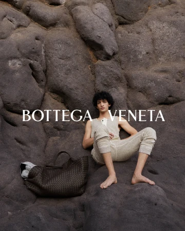 A man reclining barefoot against a rocky stone surface next to a dark woven leather tote bag for Bottega Veneta.