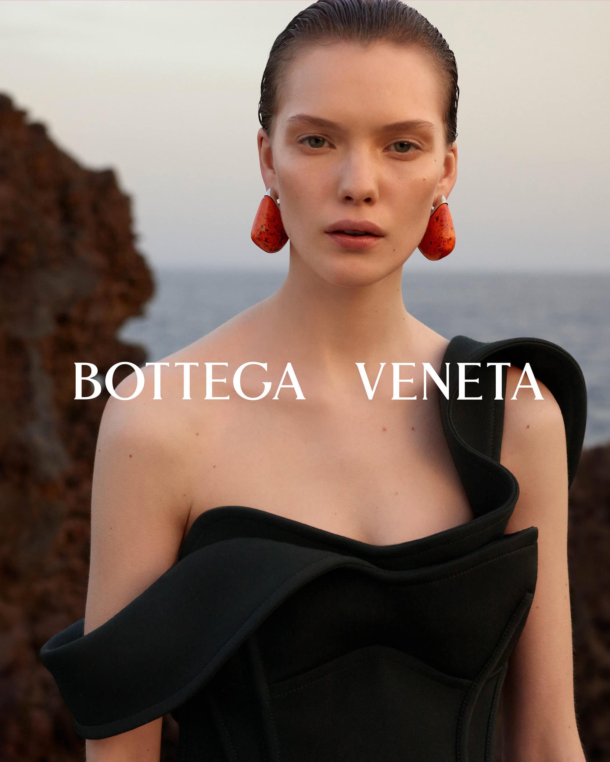 Close-up of a fair-skinned adult woman with wet hair, large orange earrings, and a black asymmetrical dress, posing outdoors by the sea.