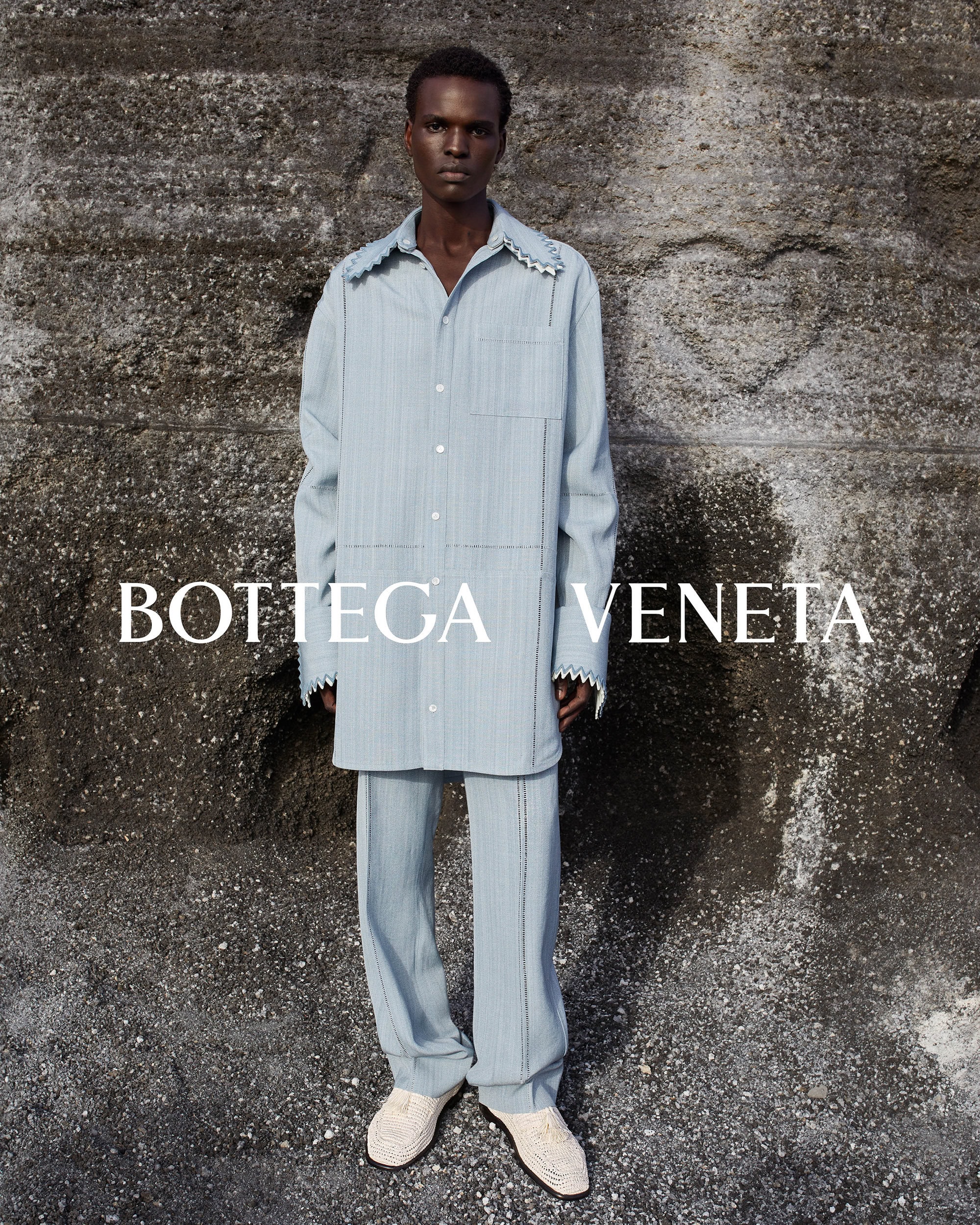 An adult male poses against a stone wall wearing a blue button-down shirt and matching pants; the brand name BOTTEGA VENETA is visible.