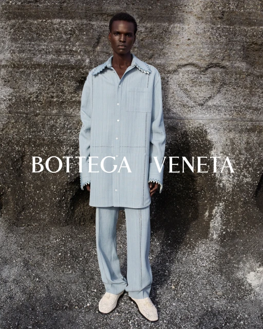 An adult male poses against a stone wall wearing a blue button-down shirt and matching pants; the brand name BOTTEGA VENETA is visible.