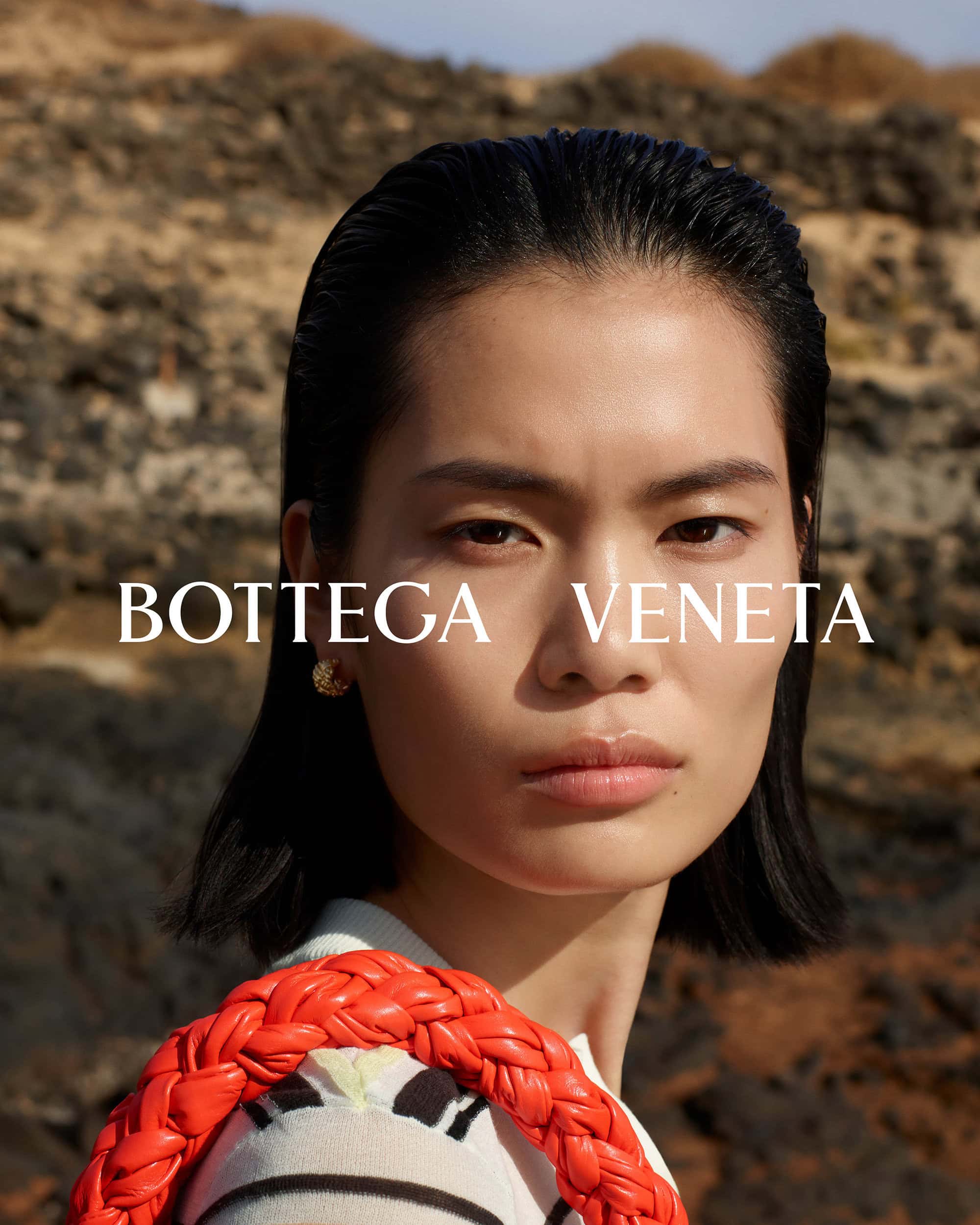 Portrait of an East Asian female model showcasing a Bottega Veneta bag, set against a textured outdoor background with natural lighting.