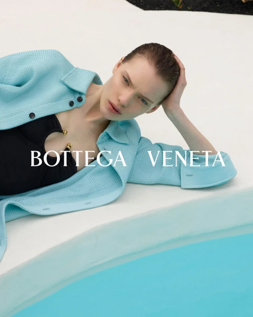 Fashion shot of a woman in a black swimsuit and a light-blue jacket lying next to a pool with the Bottega Veneta logo on the image.