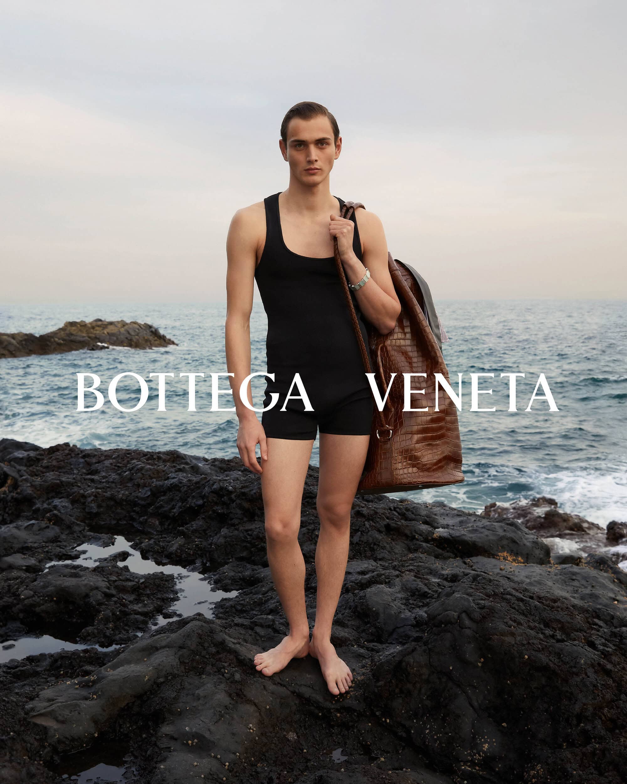 Male model wears black swimsuit with a brown leather backpack standing on rock formation in front of ocean, Bottega Veneta branding.