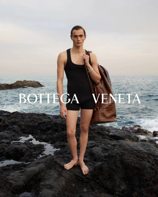 Male model wears black swimsuit with a brown leather backpack standing on rock formation in front of ocean, Bottega Veneta branding.