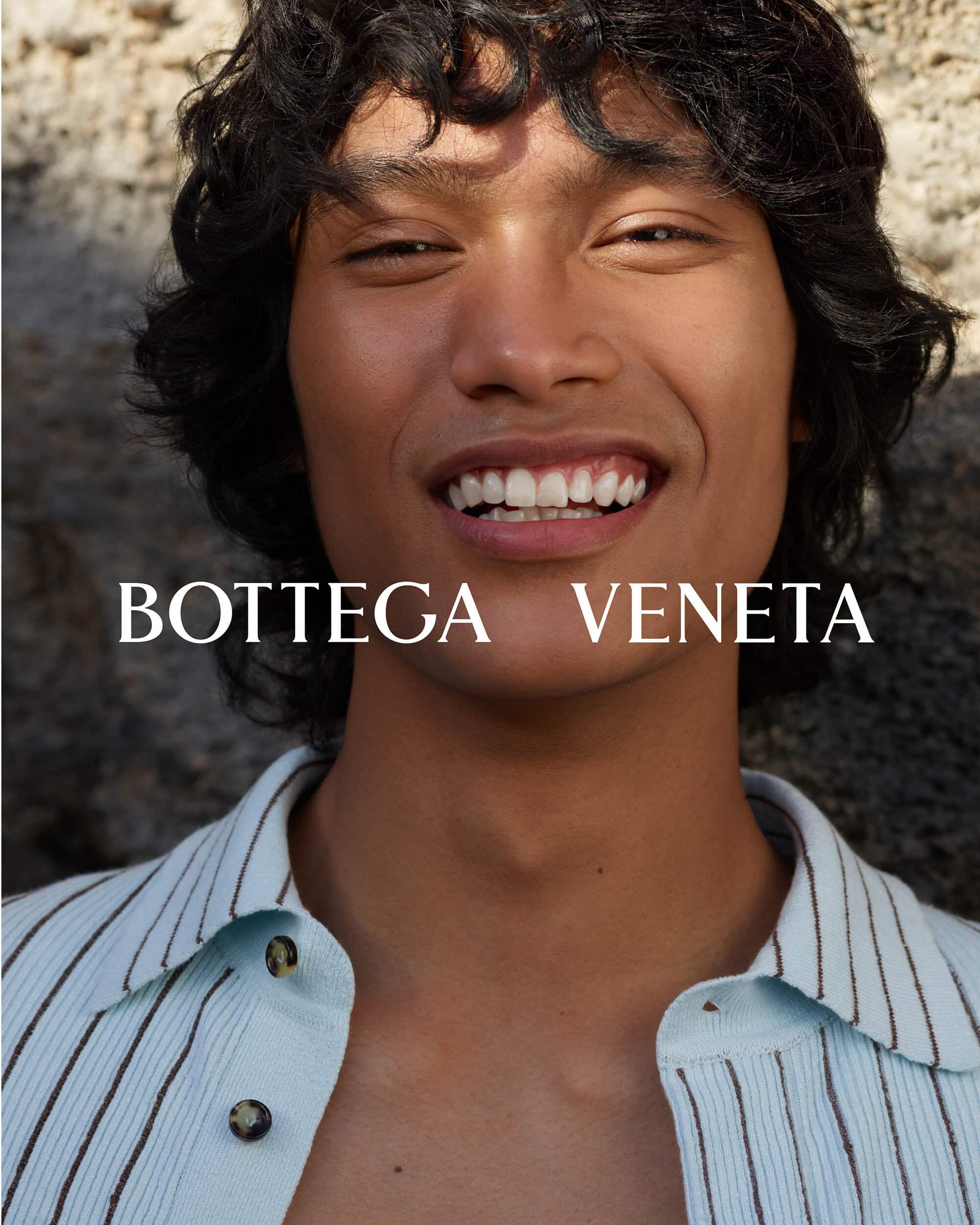 Close-up portrait of a smiling person with curly hair wearing a light blue striped shirt. The fashion brand 'Bottega Veneta' is visible.