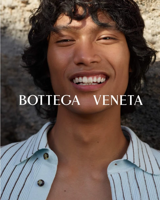 Close-up portrait of a smiling person with curly hair wearing a light blue striped shirt. The fashion brand 'Bottega Veneta' is visible.