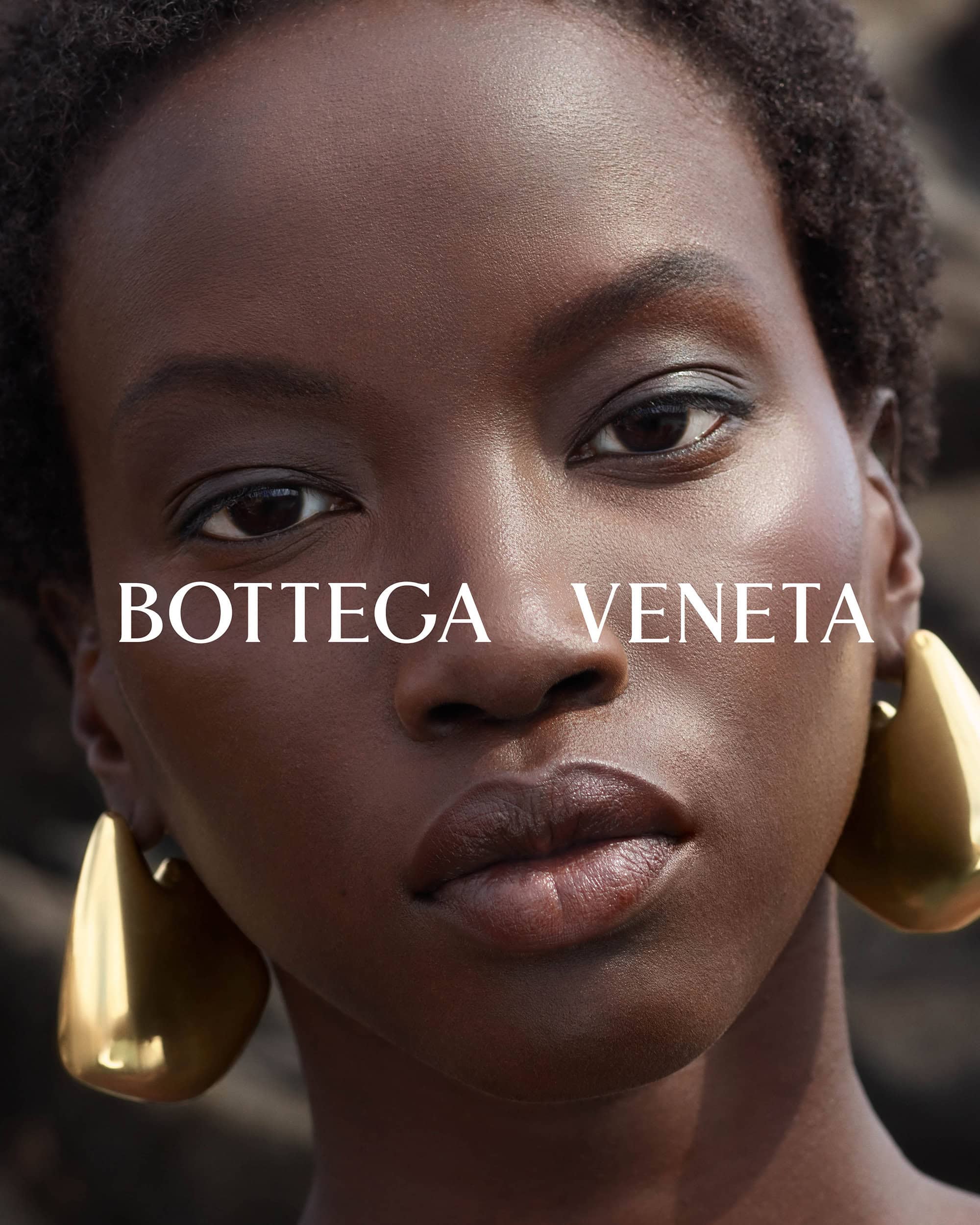 Close-up fashion shot of an adult woman of African descent with short afro, wearing gold earrings, with blurred background, and Bottega Veneta logo.