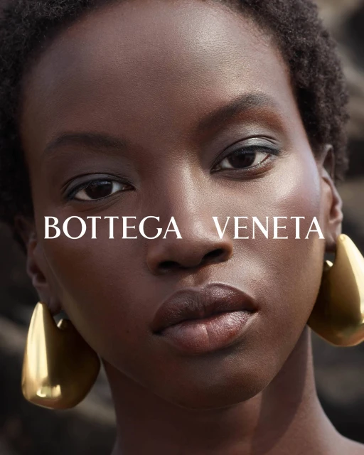 Close-up fashion shot of an adult woman of African descent with short afro, wearing gold earrings, with blurred background, and Bottega Veneta logo.