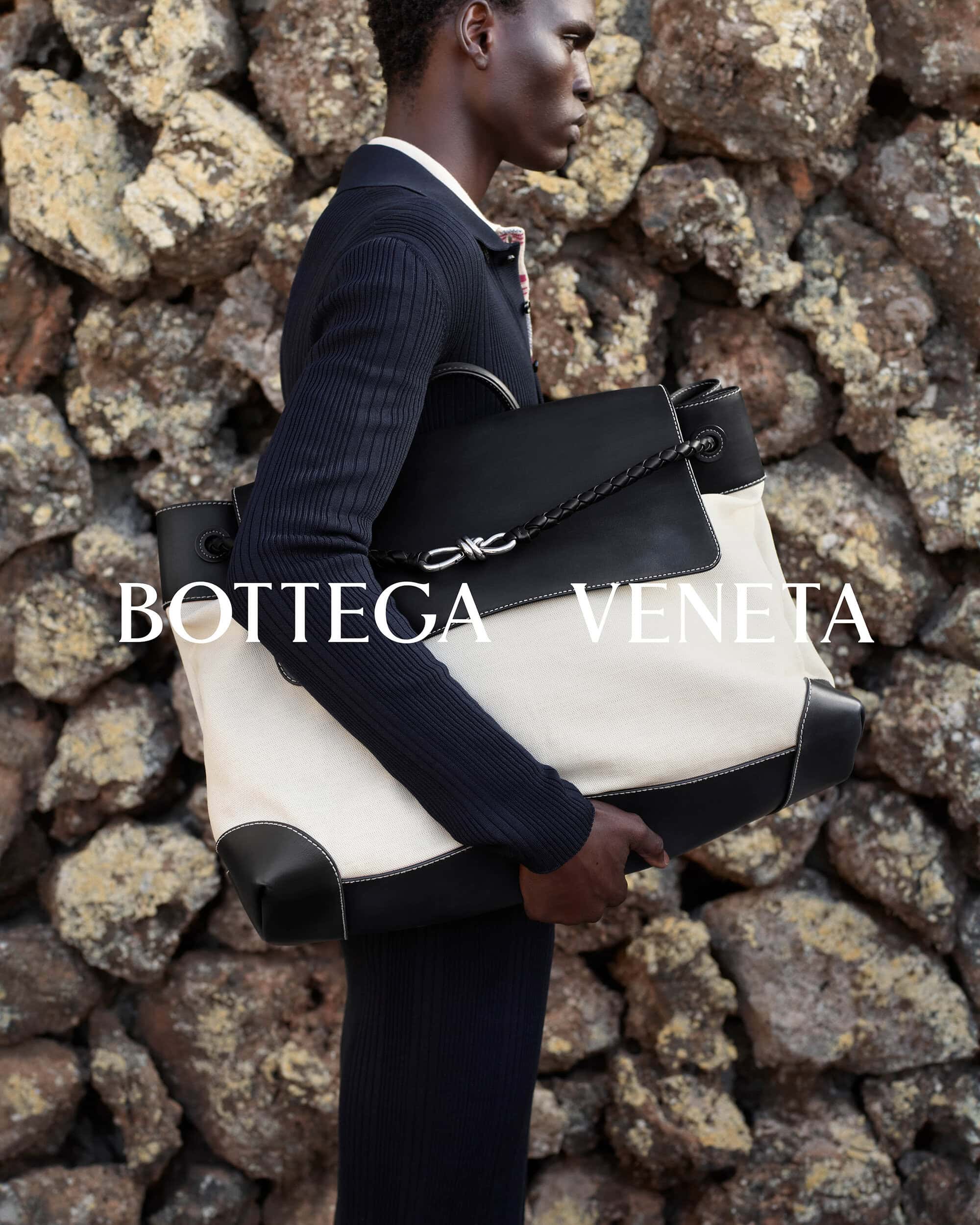An African American man models a white canvas and black leather Bottega Veneta tote bag with a stone wall background in a fashion editorial.