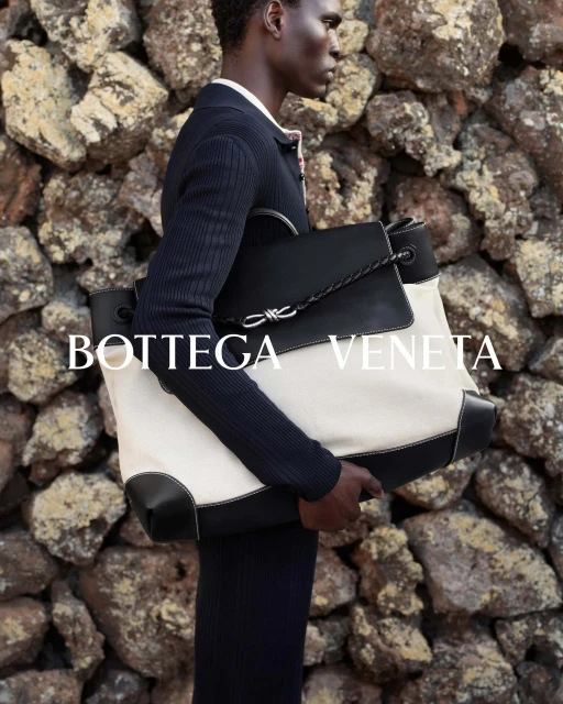 An African American man models a white canvas and black leather Bottega Veneta tote bag with a stone wall background in a fashion editorial.