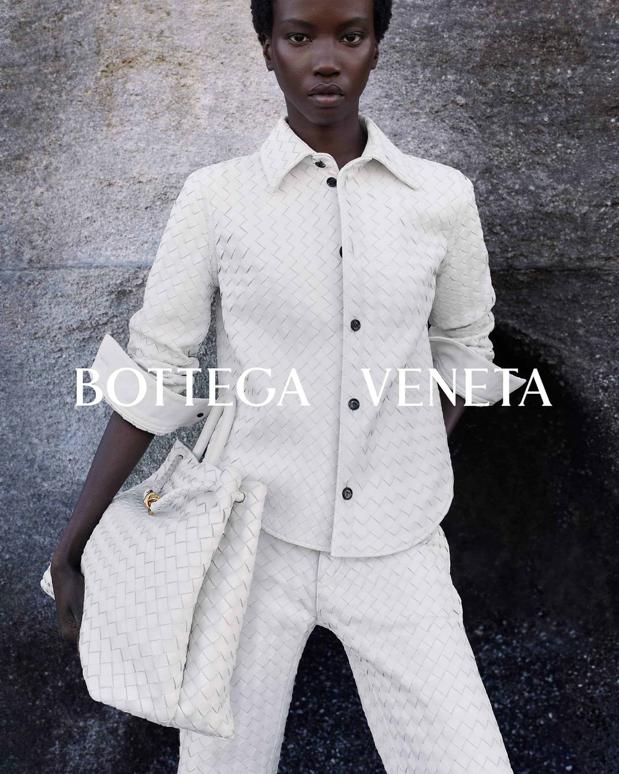 Fashion editorial shot of a Black female model in a woven cream-colored outfit and matching tote bag, posed against a textured grey background.
