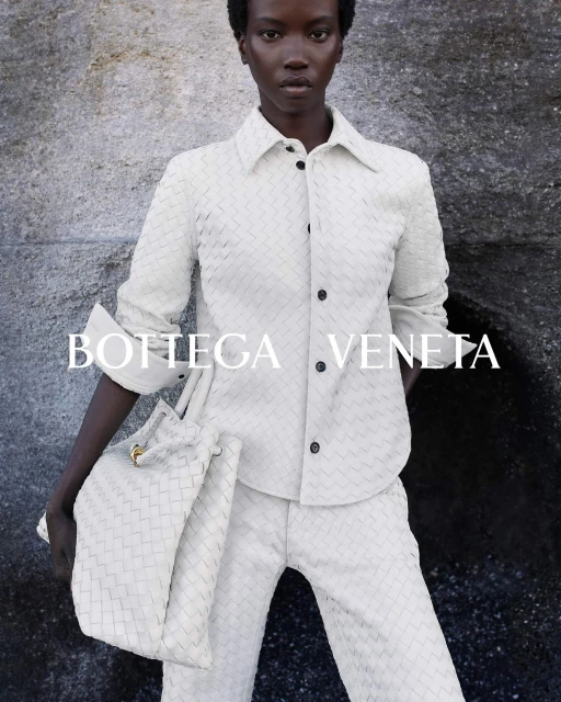 Fashion editorial shot of a Black female model in a woven cream-colored outfit and matching tote bag, posed against a textured grey background.