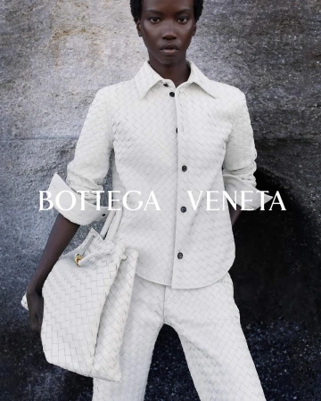 Fashion editorial shot of a Black female model in a woven cream-colored outfit and matching tote bag, posed against a textured grey background.