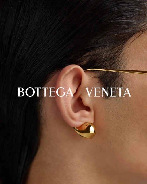 Close-up of a person's ear with a golden Bottega Veneta earring, black hair, and glasses visible against a neutral background.
