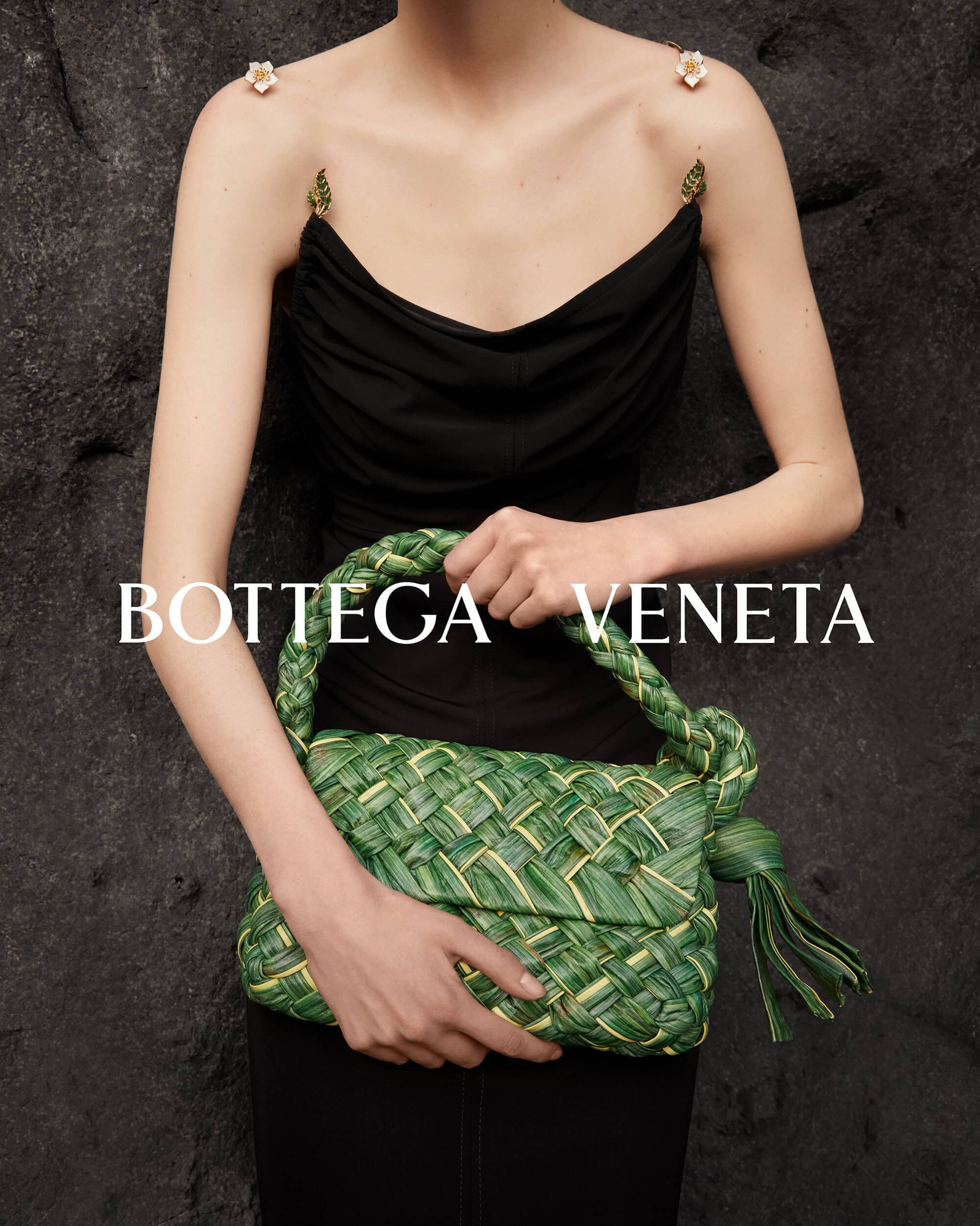 Model presents Bottega Veneta handbag made of woven green and gold material against a textured grey backdrop, showcasing fashion accessory.