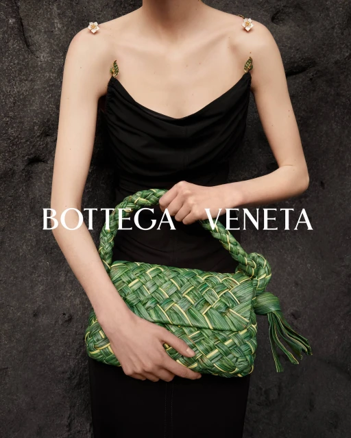 Model presents Bottega Veneta handbag made of woven green and gold material against a textured grey backdrop, showcasing fashion accessory.