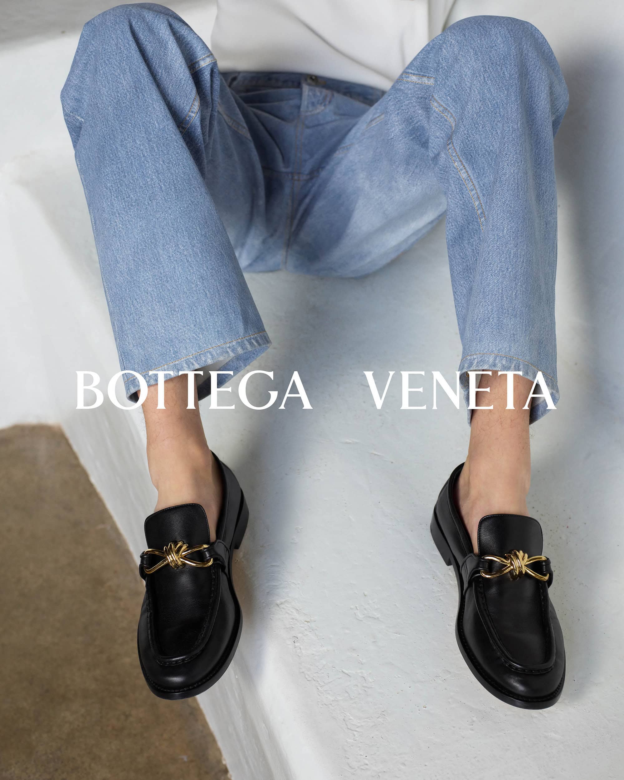 Person wearing light wash blue jeans and black leather loafers with gold hardware seated on a light surface.