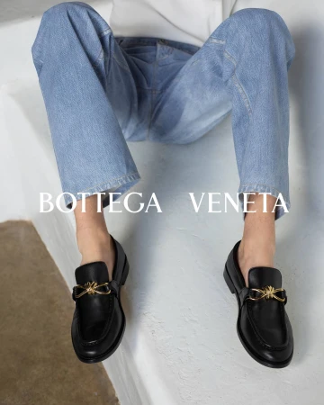 Person wearing light wash blue jeans and black leather loafers with gold hardware seated on a light surface.