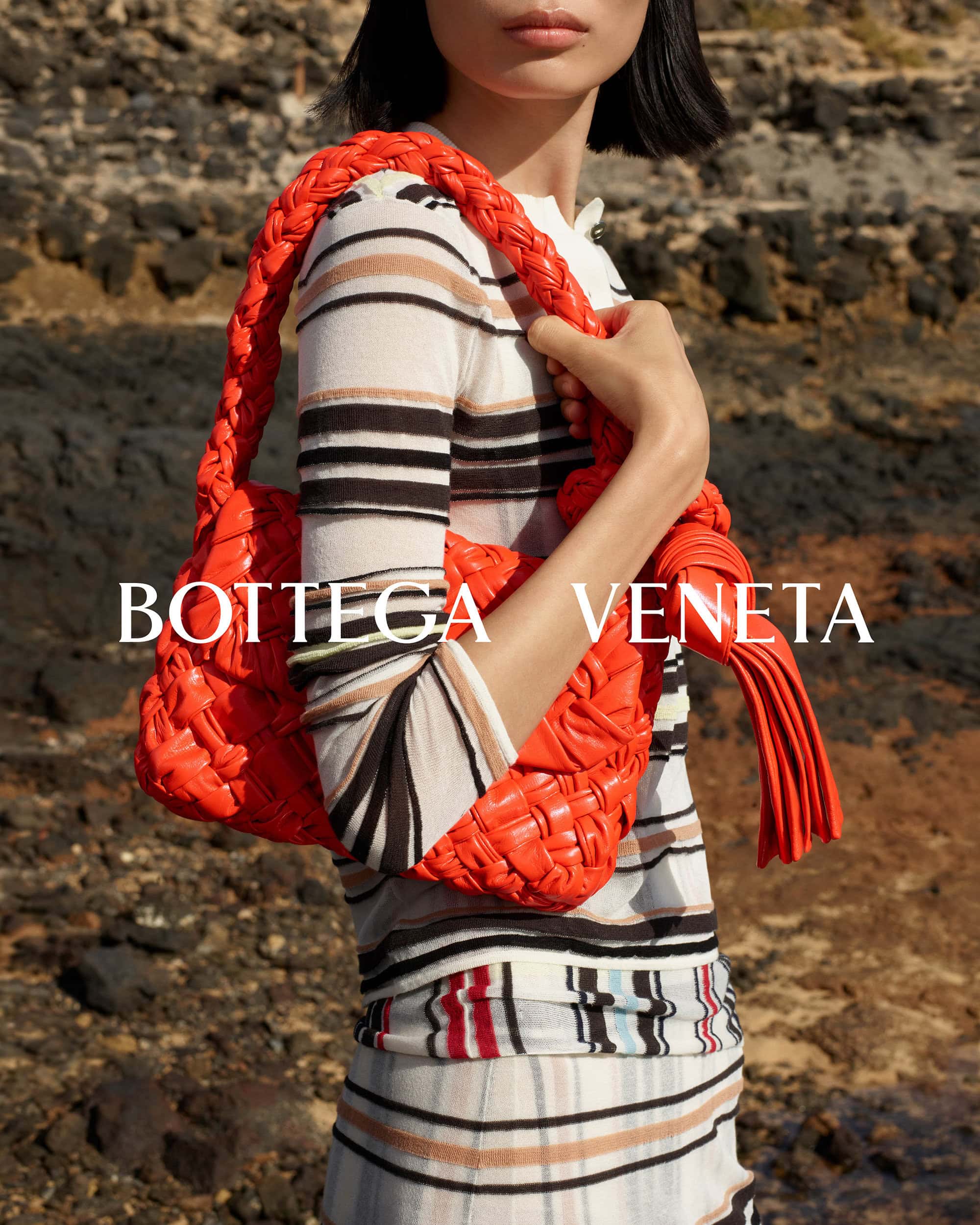 Model holding a bright orange woven leather handbag wearing a striped knit top against a rocky background for Bottega Veneta