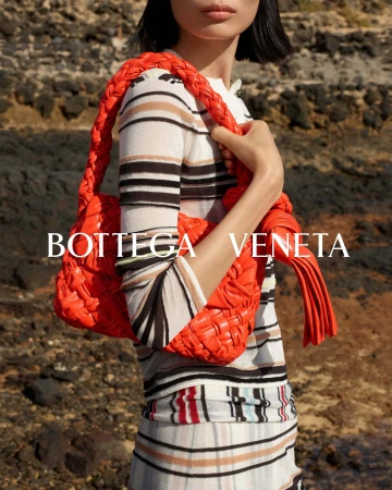 Model holding a bright orange woven leather handbag wearing a striped knit top against a rocky background for Bottega Veneta