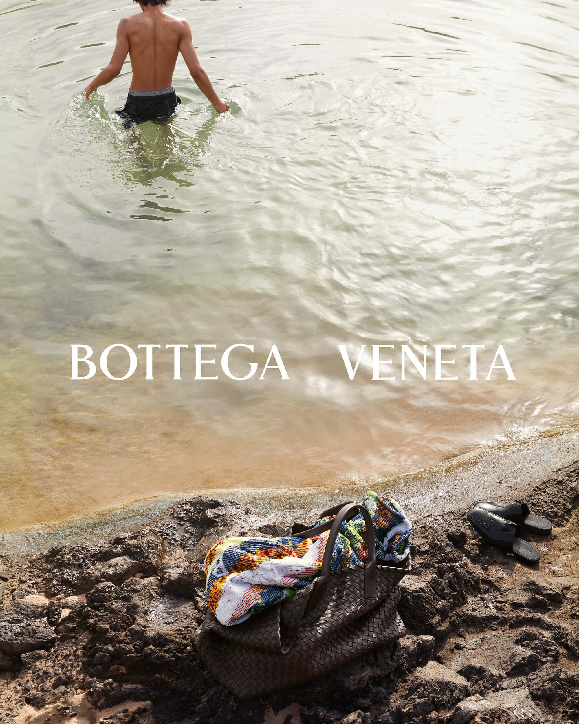 A person is seen wading in water toward the beach. A woven Bottega Veneta handbag and sandals rest on the rocky sand.