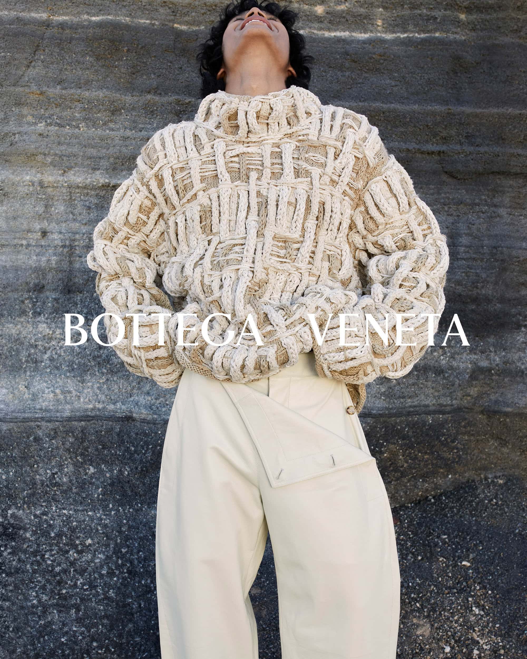 Fashion shot of a model wearing a chunky knit sweater and cream-colored pants against a textured rock background, Bottega Veneta brand.
