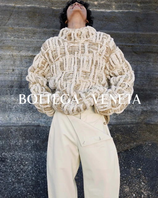 Fashion shot of a model wearing a chunky knit sweater and cream-colored pants against a textured rock background, Bottega Veneta brand.