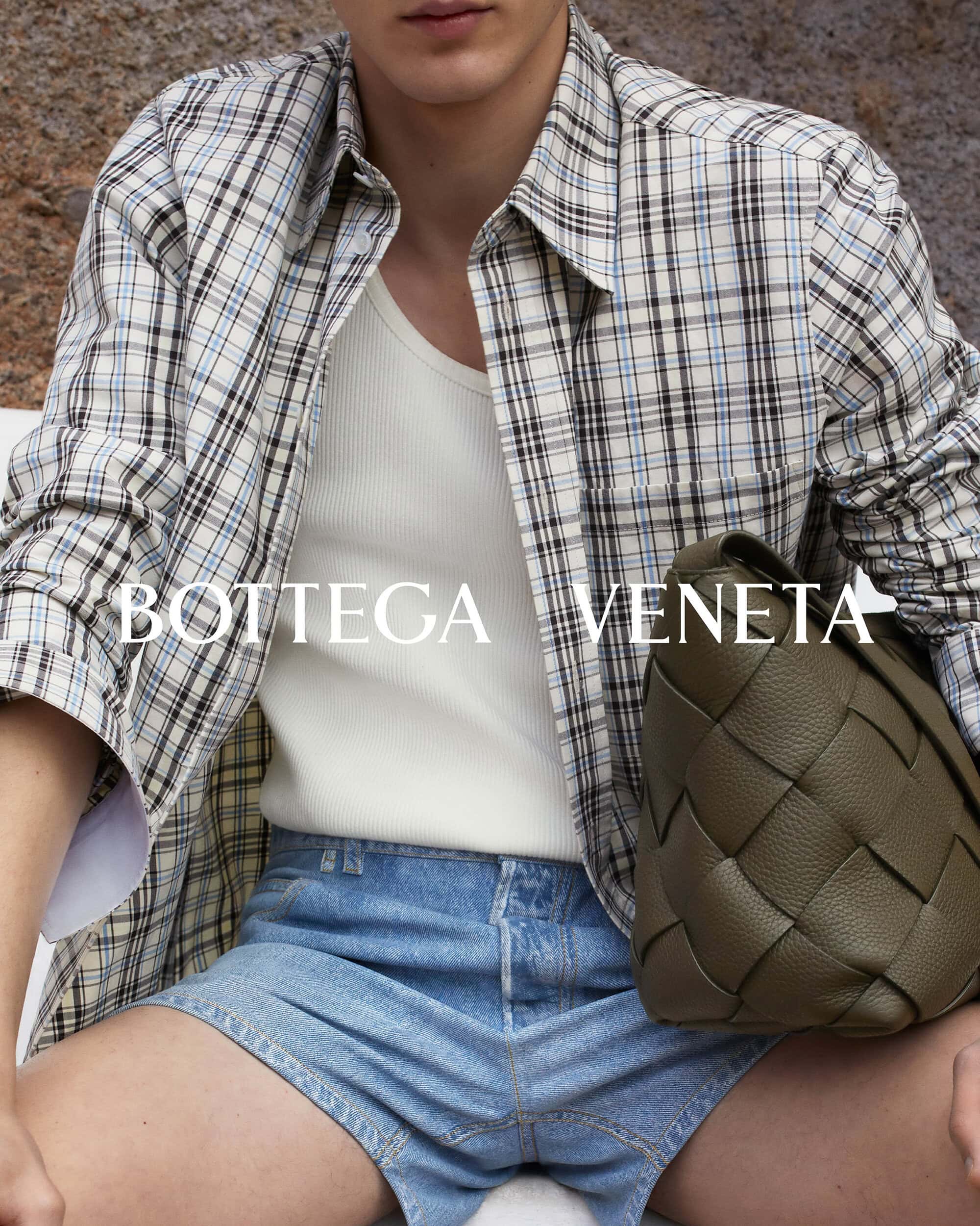 A person wearing a plaid shirt, tank top, and denim shorts is shown indoors, posing with a brown Bottega Veneta woven bag.