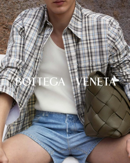 A person wearing a plaid shirt, tank top, and denim shorts is shown indoors, posing with a brown Bottega Veneta woven bag.