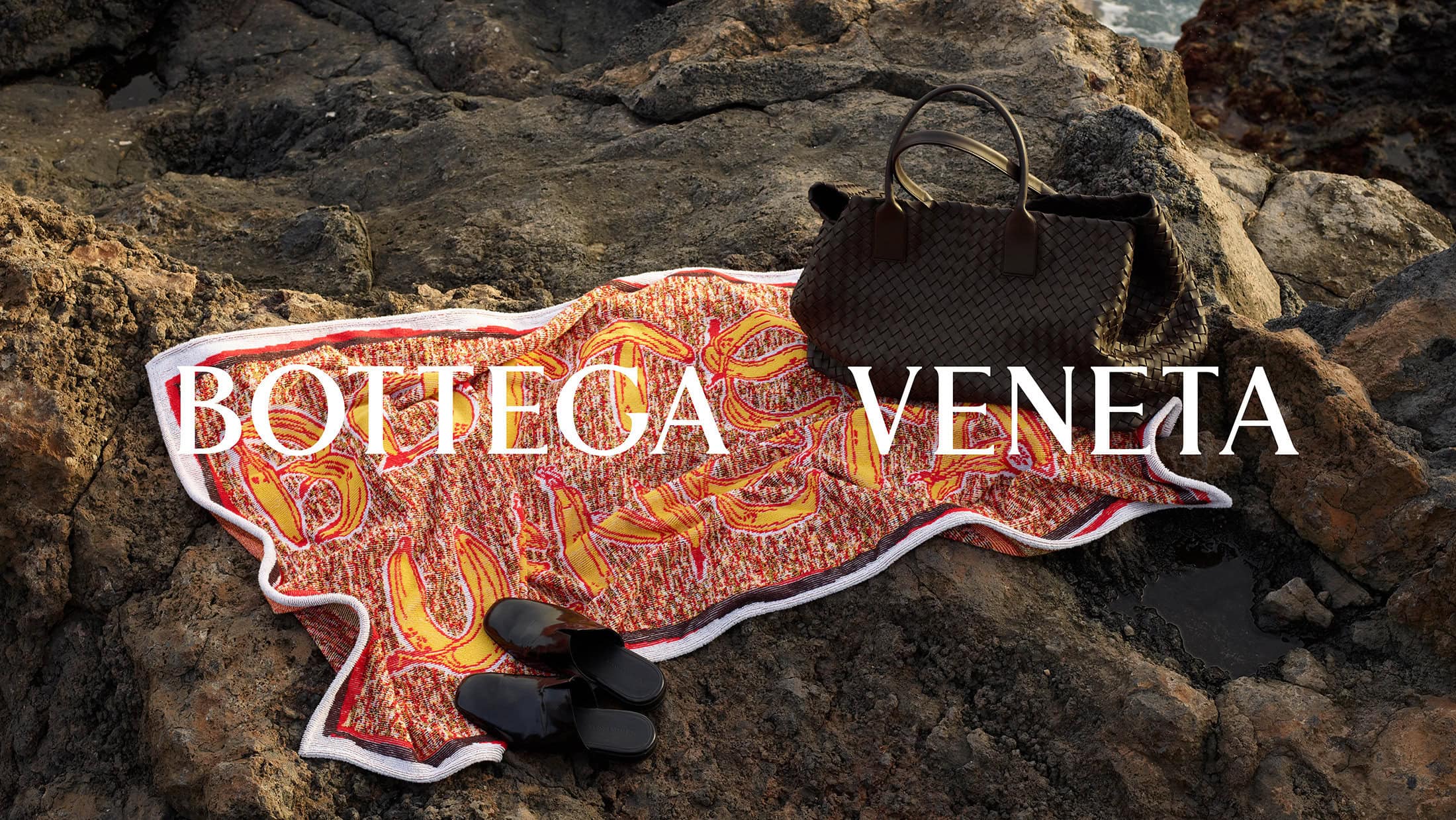 Still life of a Bottega Veneta handbag and sandals on a graphic towel, placed on rocks near the ocean, showcasing a beach-ready fashion scene.