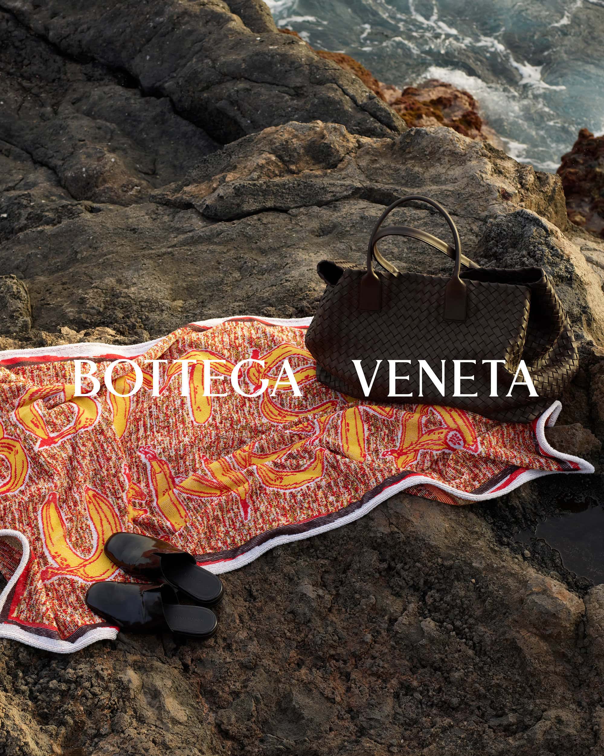 Bottega Veneta leather handbag, patterned towel, and black slip-on shoes arranged on wet rocky coast by crashing ocean waves.