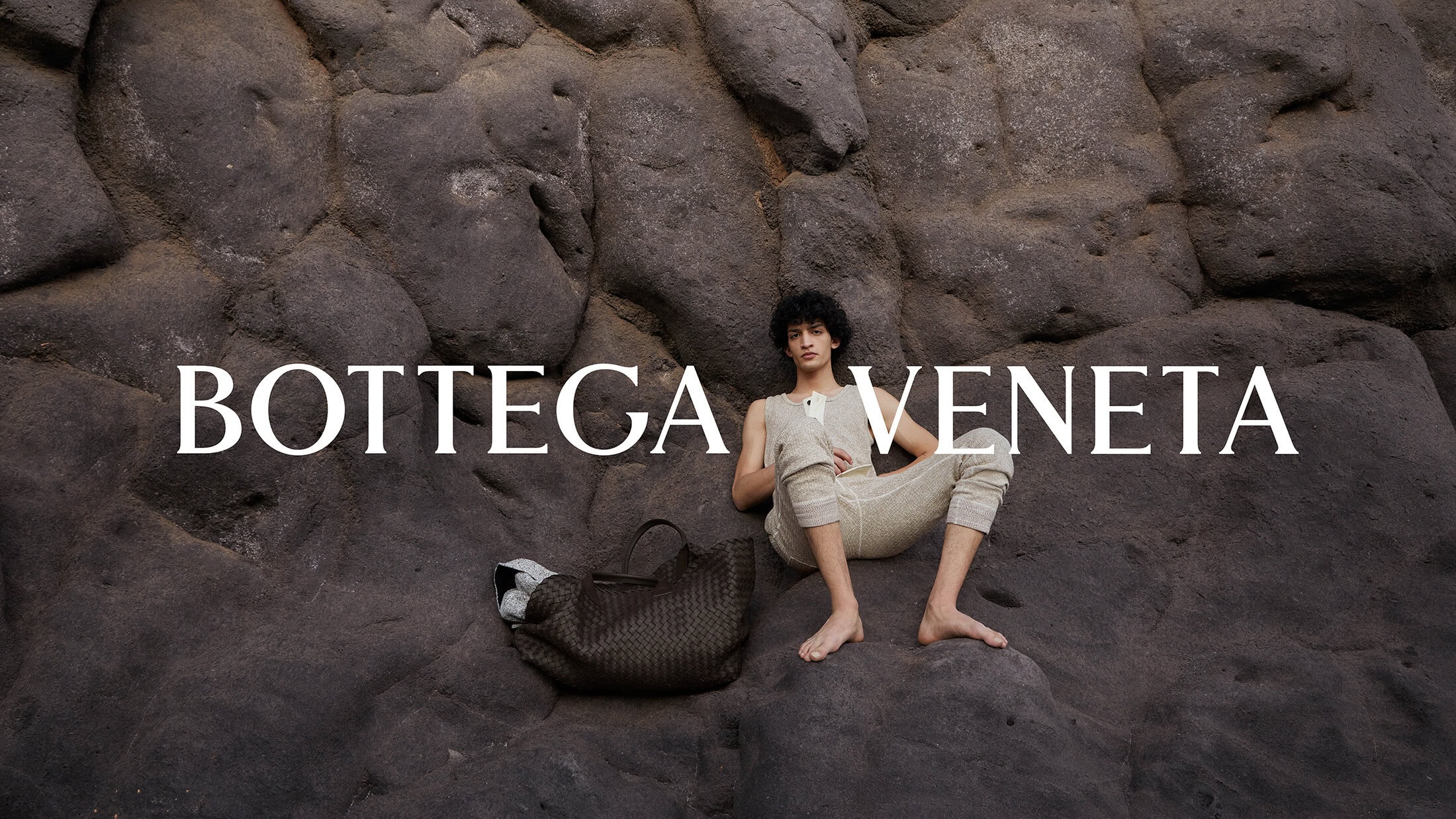 A male model in an off-white knit outfit poses on a textured grey-black rock with a woven tote bag in an outdoor fashion editorial.