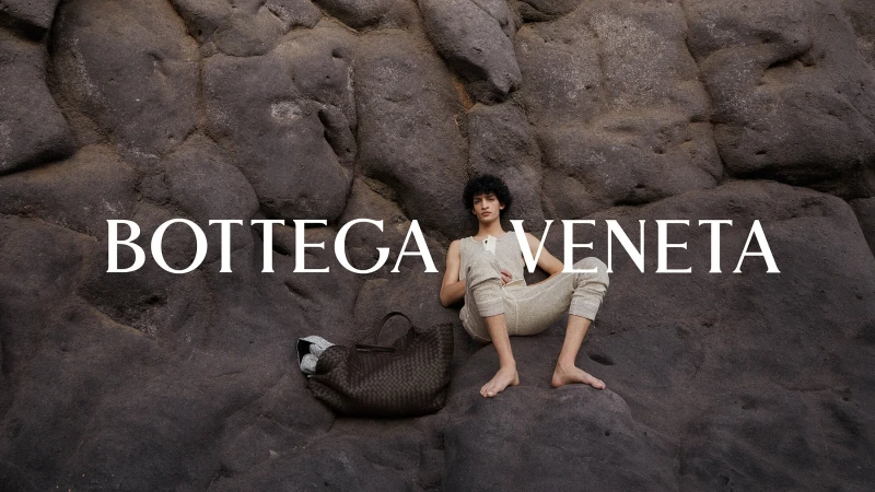 A male model in an off-white knit outfit poses on a textured grey-black rock with a woven tote bag in an outdoor fashion editorial.