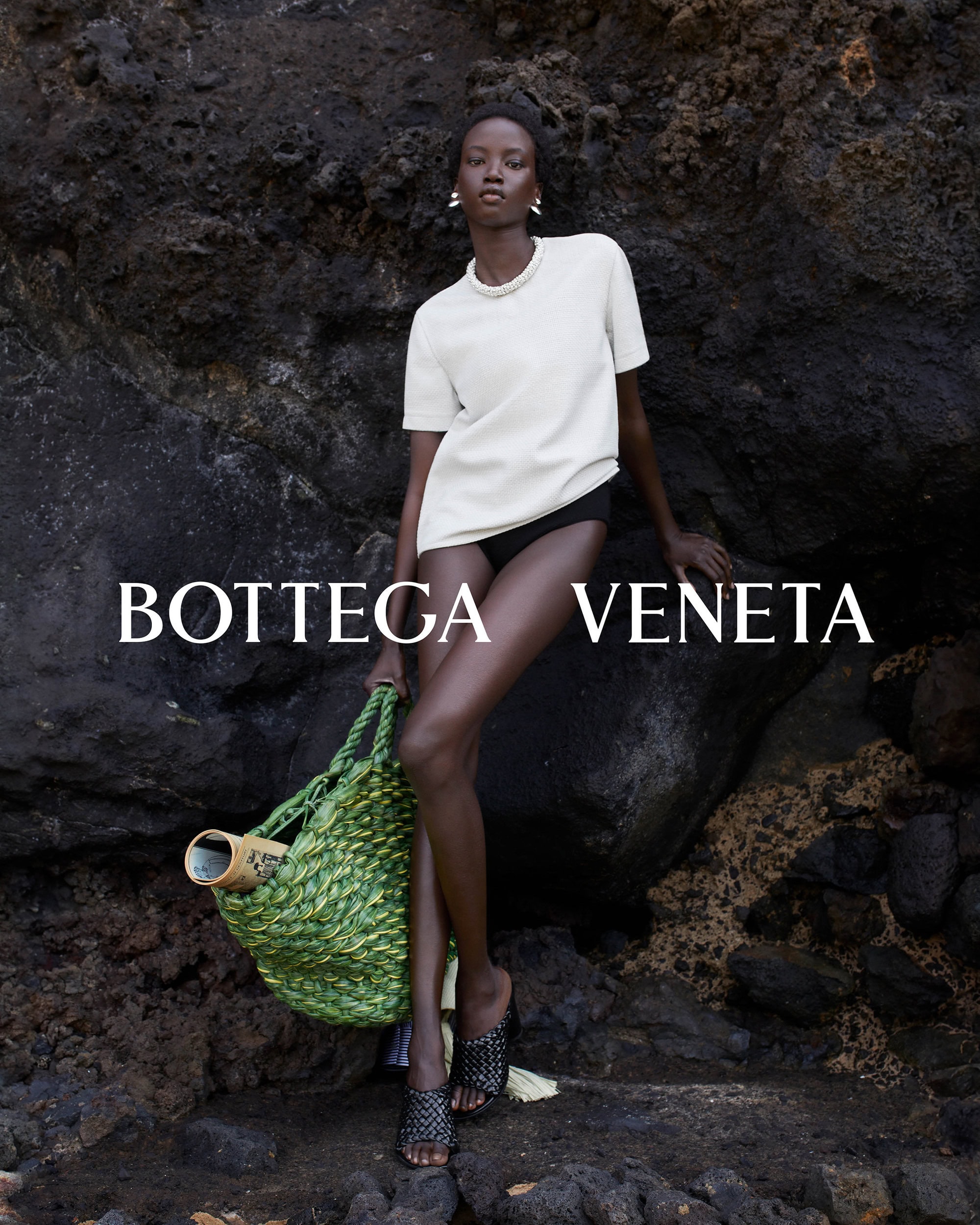 A woman in a cream top and dark bottoms poses with a green woven bag against a textured dark rock background in a fashion shot.
