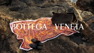 Bottega Veneta leather handbag and patterned beach towel laid out on dark rocky coastal ground.