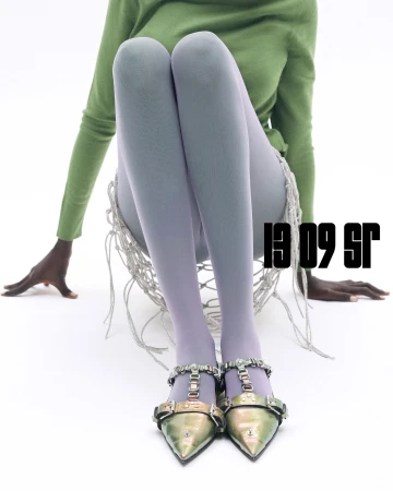 Model seated on a white floor wearing a green top, light grey tights, and metallic embellished shoes against a seamless white background.