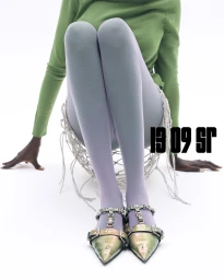 Model seated on a white floor wearing a green top, light grey tights, and metallic embellished shoes against a seamless white background.