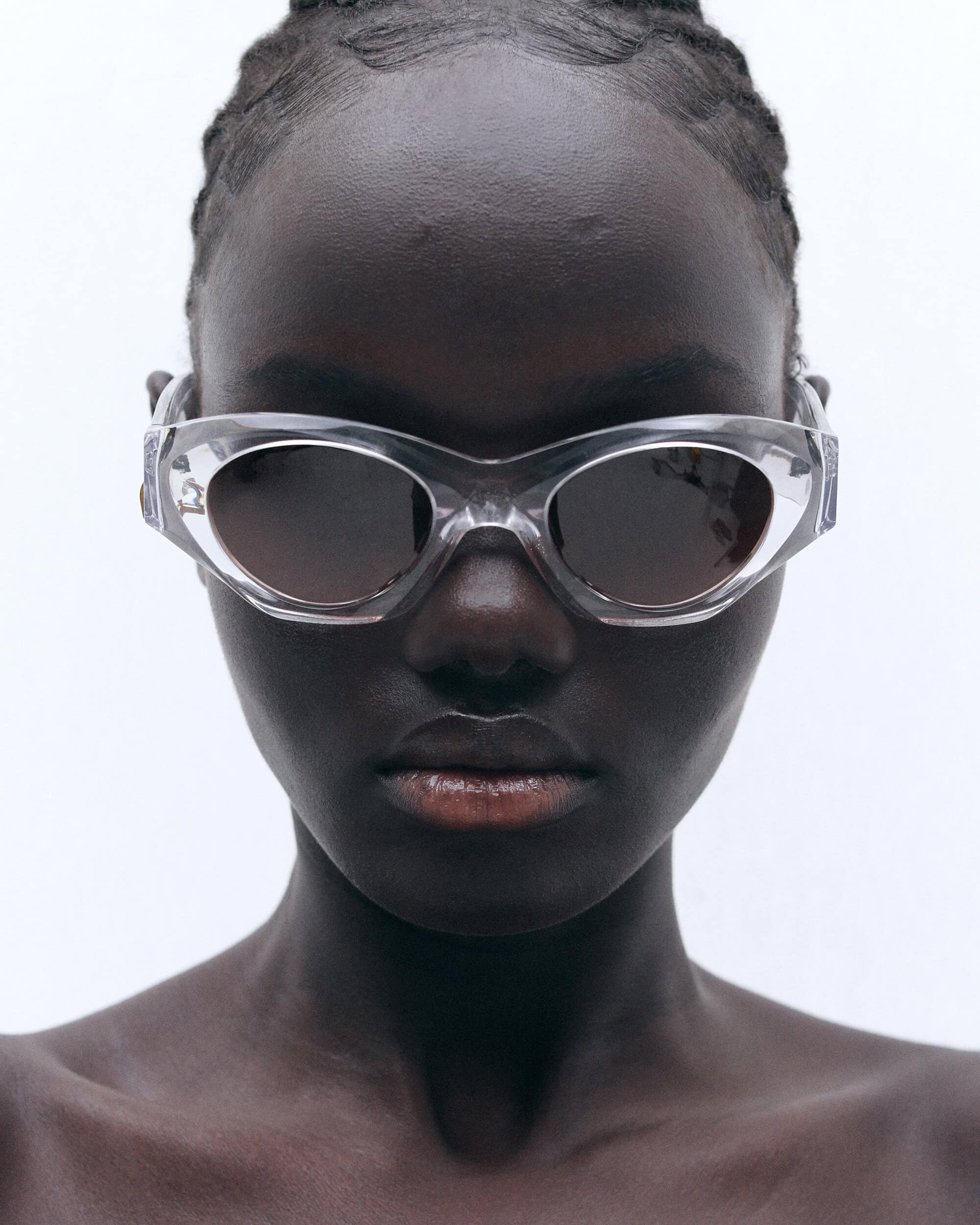 Close-up portrait of a woman with dark skin wearing clear-framed sunglasses, showcasing fashion and beauty against a bright background.