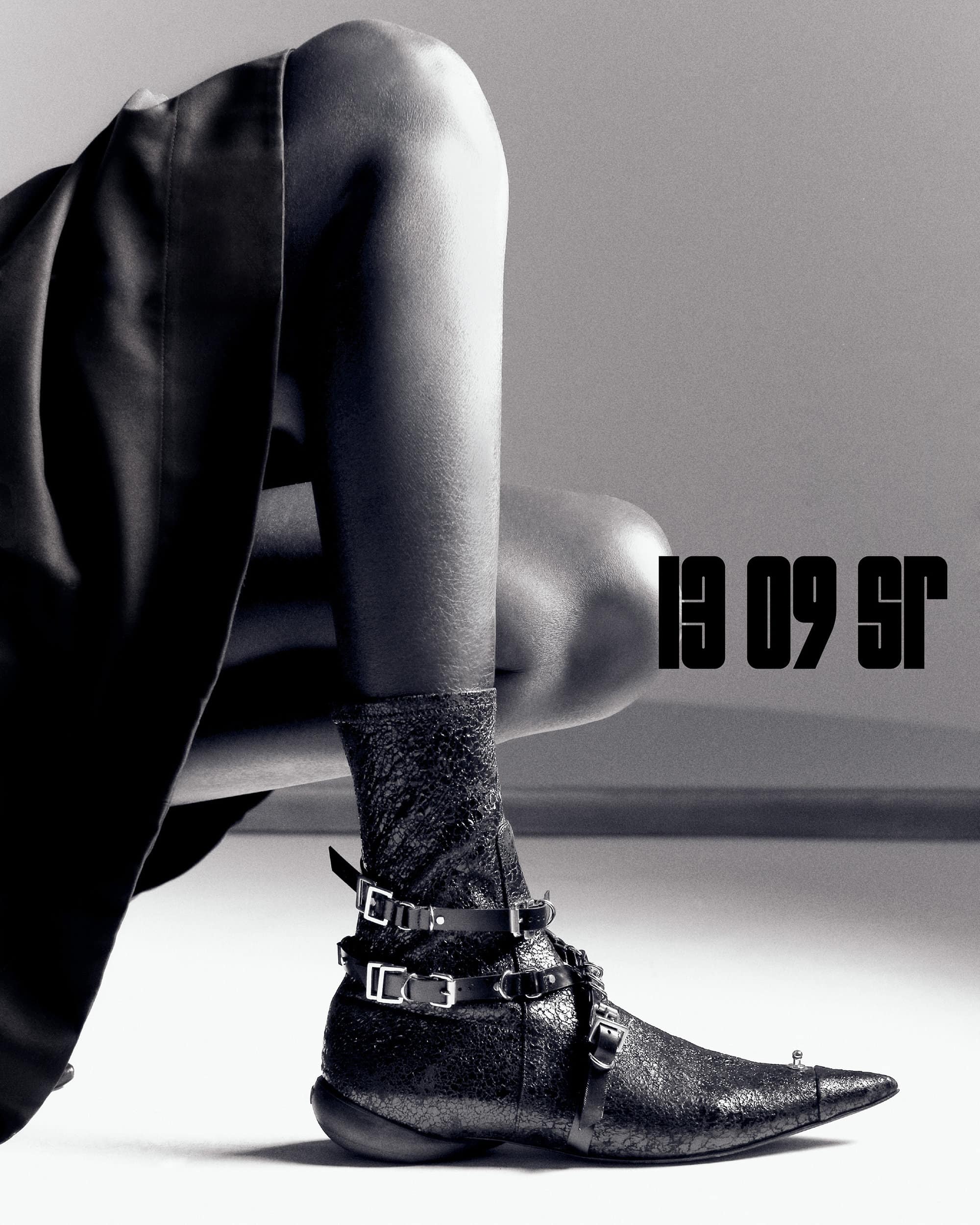Close-up of a woman's leg wearing a decorative black ankle boot with straps, buckles, and a pointed toe against a grey studio backdrop.